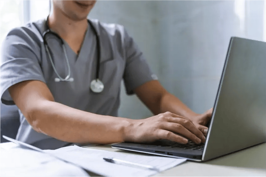 Un profesional de la salud usando una laptop para trabajar en un escritorio, con un estetoscopio alrededor del cuello y documentos y bolígrafos al lado