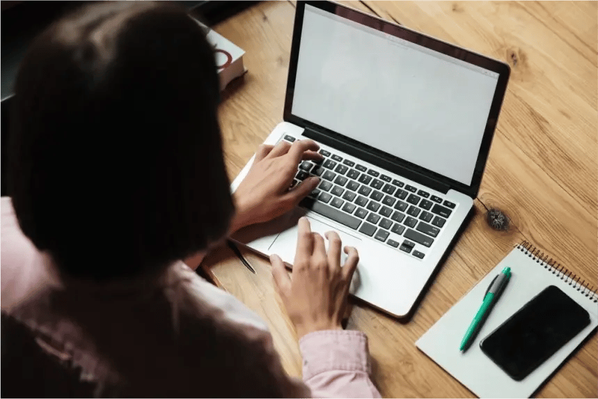 Una persona trabajando en una laptop para trabajar sobre un escritorio de madera, con un cuaderno, un bolígrafo verde y un teléfono al lado, enfocándose en una pantalla en blanco-1